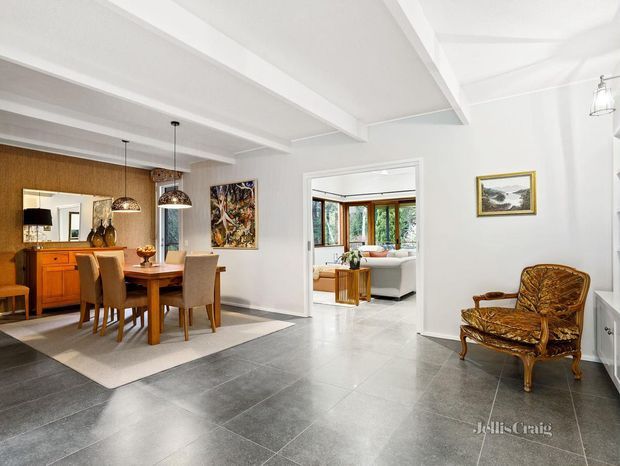 Bright open-plan interior showing a dining area in the foreground, a living room visible through a wide doorway, and a sitting chair in the corner. The space has large dark grey floor tiles, white walls and exposed white ceiling beams, warm timber dining furniture and cabinetry, pendant lighting, and abundant natural light.