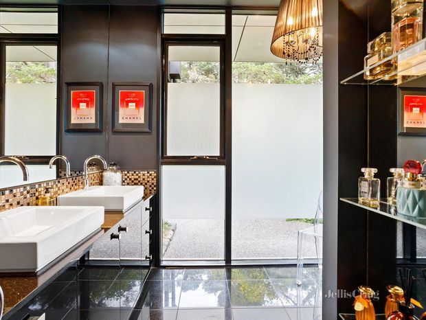 A sophisticated, high-contrast bathroom with two white vessel basins on dark cabinetry, polished black floor tiles, dark-framed frosted glazing, and a mosaic tile splashback in cream, tan, brown, and black. Open black shelving displays glassware and decorative bottles, while a large warm pendant light adds a luxurious accent. The room opens toward a bright outdoor area through full-height glazed doors.