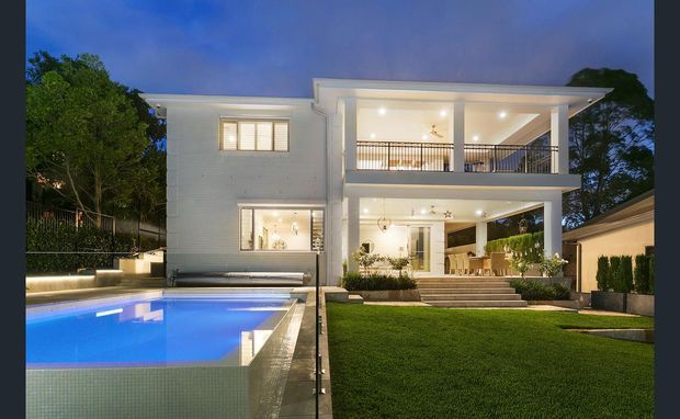 Dusk exterior view of a large two-storey contemporary home with a crisp white facade, broad covered upper and lower verandas, illuminated interiors, landscaped lawn, and a rectangular swimming pool with glass fencing in the foreground.
