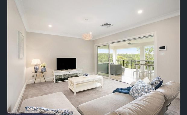 Bright contemporary living room with a large pale sectional sofa, white coffee table and TV console, neutral carpet, and recessed ceiling lighting. Wide sliding glass doors open onto a covered balcony or terrace with outdoor seating and greenery beyond.