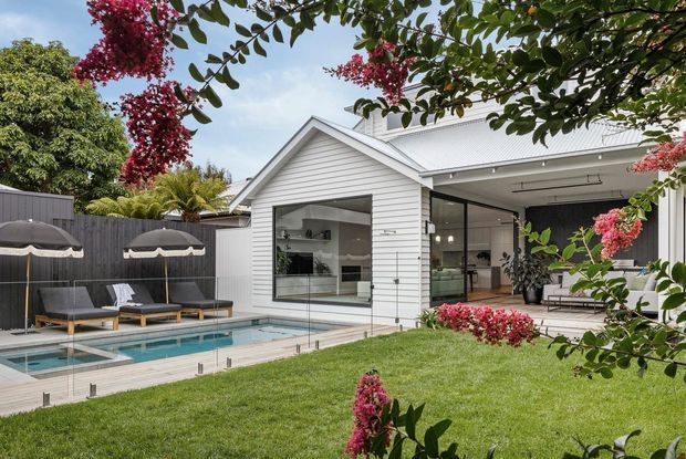 A landscaped residential backyard with a white weatherboard house, pitched gable roof, large glazed openings, and a covered outdoor living area. The space includes a rectangular swimming pool with glass fencing, paved pool terrace, sun loungers, umbrellas, lawn, and flowering garden planting. The open kitchen and living areas are visible through the glazing.