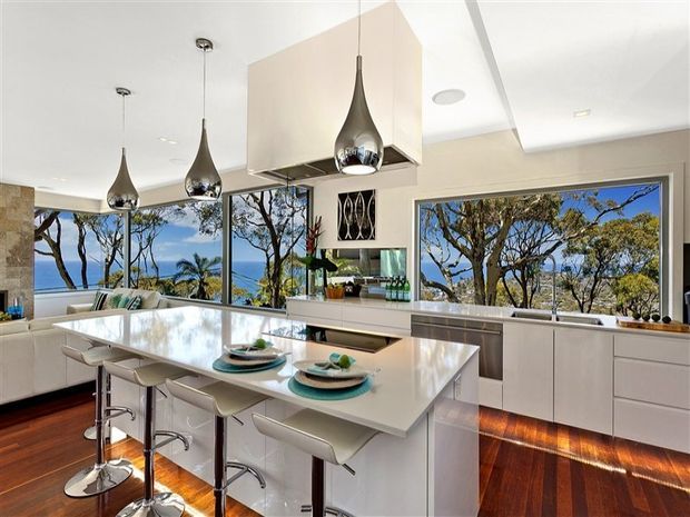 Bright contemporary kitchen with a large white island, integrated seating for four, white cabinetry, stainless-steel appliances, and sculptural pendant lights. Floor-to-ceiling glazing provides expansive views of coastal vegetation and blue water, while dark timber flooring and under-cabinet lighting add warmth.