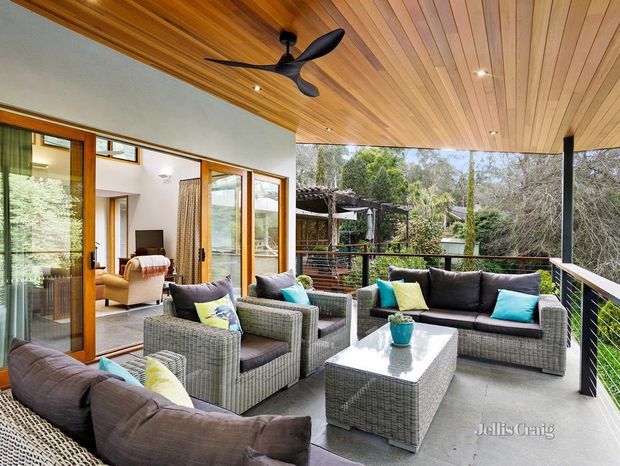 Covered outdoor terrace with a timber-lined ceiling, black ceiling fan, glass doors opening to an interior living room, and a large outdoor seating arrangement overlooking trees. The space features woven grey sofas and armchairs, a matching coffee table, bright cushions, dark metal posts, and a black balustrade.