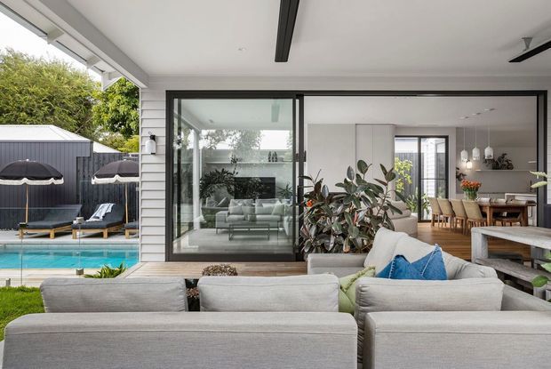 A bright contemporary indoor-outdoor living area opening onto a swimming pool and landscaped yard. The foreground features a large light-grey outdoor sectional with blue accent cushions, while black-framed sliding glazing connects to an interior living room and dining area. White horizontal weatherboard cladding, a covered patio, ceiling fans, timber decking, and abundant greenery create a relaxed residential setting.