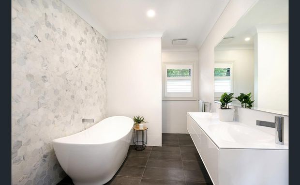 Bright contemporary bathroom featuring a freestanding white bathtub, a large white double vanity with integrated basins, dark grey tiled flooring, a pale patterned feature wall, mirrored cabinetry, and plantation shutters. The room has a clean, refined, high-contrast finish with abundant natural light.