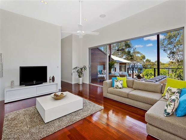 Bright contemporary living room with polished dark timber flooring, white walls and ceiling, a large white media console, low white coffee table, and neutral upholstered sofas. Full-height glazing opens the room visually to a leafy outdoor balcony or terrace, with a ceiling fan and abundant natural light.