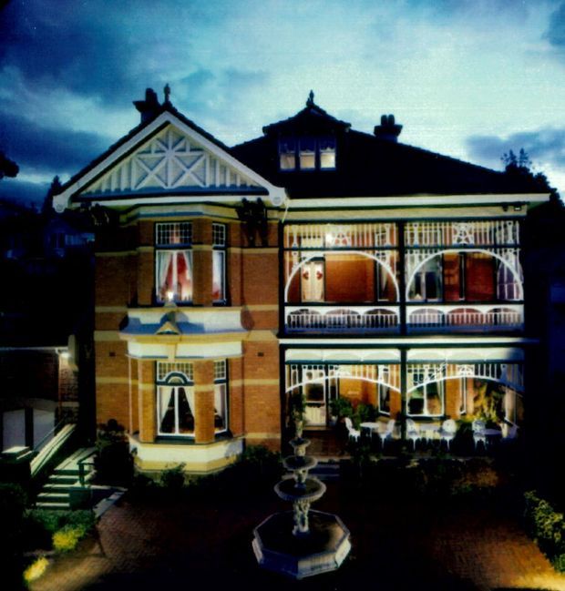 A grand multi-storey historic residence photographed at dusk, featuring a steep gabled roof, ornate timber fretwork, decorative verandahs and balconies, contrasting brickwork, bay windows, and a formal courtyard with a central fountain.