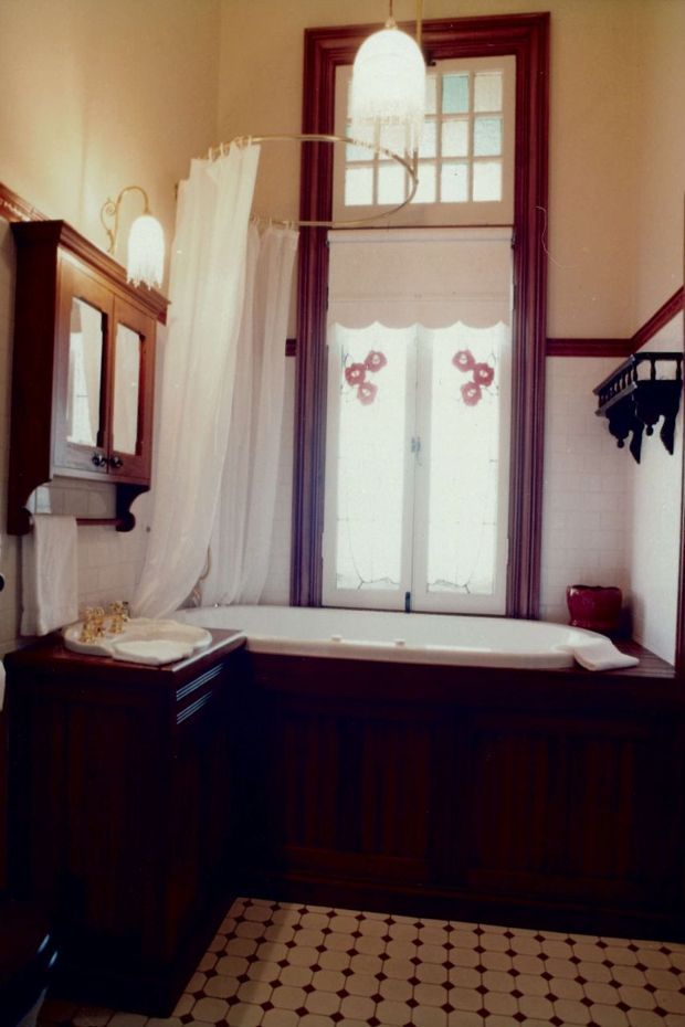 A traditional bathroom featuring a freestanding bathtub enclosed in dark timber-panelled cabinetry, a tall leadlight-style window with deep burgundy timber trim, white tiled lower walls, a vintage mirrored wall cabinet, and a sheer white shower curtain. Decorative pendant lighting and patterned floor tiles reinforce the heritage character.