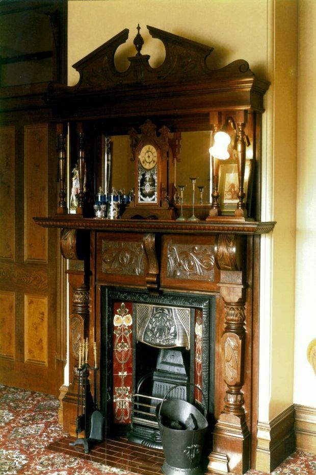 A richly detailed historic interior centred on an ornate dark timber fireplace and mantel. The carved mantel includes a mirrored overmantel, decorative cresting, turned columns, and a prominent antique clock, with patterned fireplace tiles, traditional fireplace tools, and floral patterned carpet. A paneled timber door and warm cream walls reinforce the heritage character.