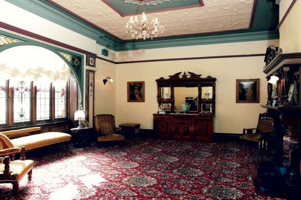 An ornate, traditionally styled sitting room or living room with a richly patterned wall-to-wall rug, dark carved timber furniture, leaded and stained-glass windows, decorative wall art, a fireplace mantel, and an elaborately painted coffered ceiling with chandelier. The room has a formal, historic character and abundant natural light.