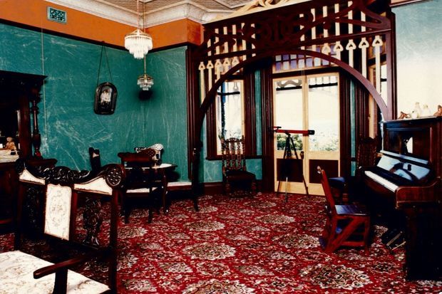 Ornate historic-style living room or salon with dark carved timber furniture, a rich red patterned carpet, deep teal walls, decorative plaster cornices, hanging chandeliers, an arched timber room divider, tall glazed doors, and an upright piano.
