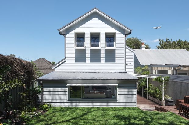 Front exterior view of a compact two-storey home with white horizontal weatherboard cladding, a prominent front gable, corrugated metal roofing, and symmetrically arranged upper-level windows. The lower level has broad glazing opening toward a landscaped garden, with a timber deck and neighbouring homes visible around it.