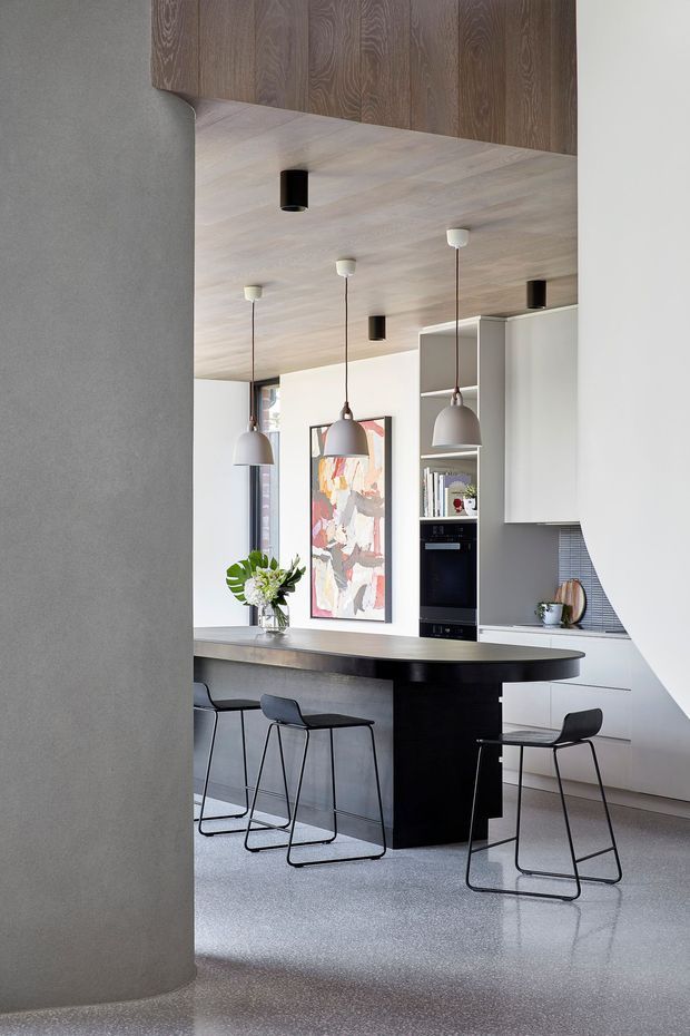 A contemporary kitchen viewed through a curved opening, with a large dark island and integrated seating, pale wood ceiling, dark wood overhead cabinetry, white cabinetry, built-in black oven, pendant lighting, and a terrazzo-like grey floor. An abstract artwork and small plant arrangement add restrained colour.