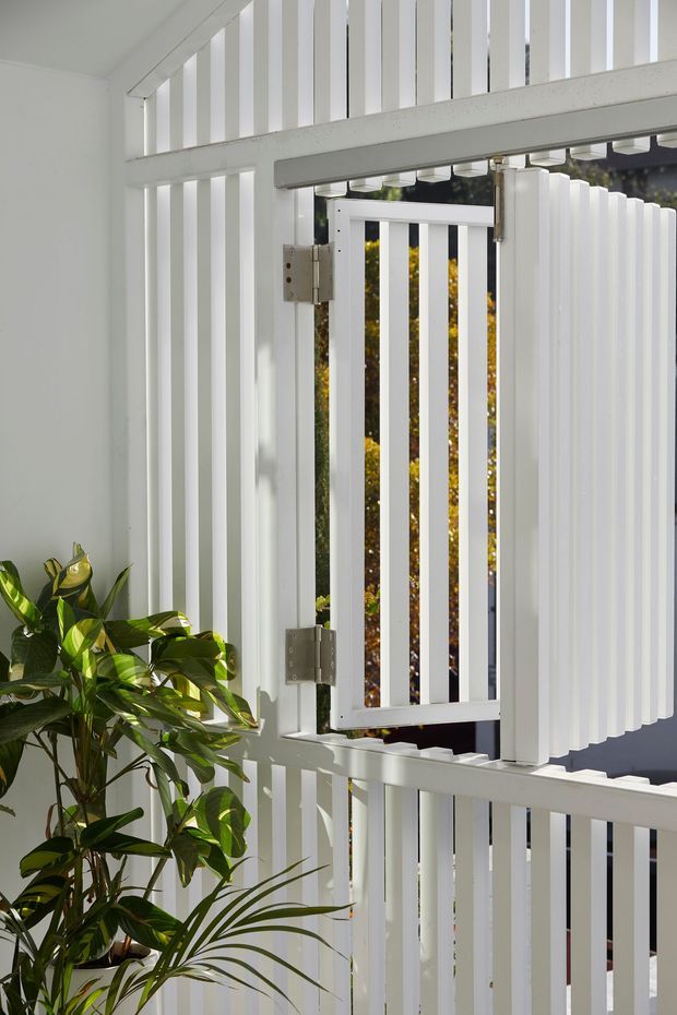 Close-up view of a white painted slatted gate and surrounding vertical privacy screen, with visible metal hinges and gate hardware. Bright natural light filters through the open gate, while leafy potted plants occupy the lower left foreground. The image focuses on an exterior architectural enclosure rather than an interior room.
