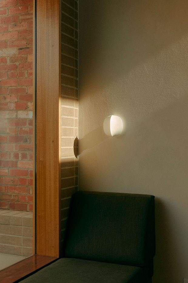 A warmly lit interior corner featuring a dark upholstered armchair beside a timber-framed opening. Exposed red brick and dark brickwork contrast with a smooth warm beige plaster wall, while a small round wall light casts a soft glow and shadow. The image emphasizes tactile materials, restrained furnishings, and intimate atmospheric lighting.