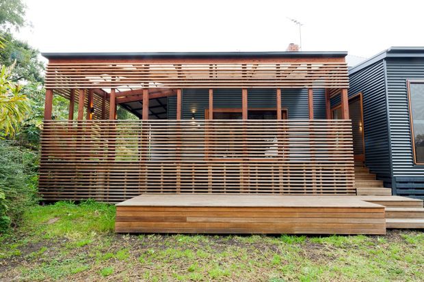 Exterior view of a compact contemporary home with a broad timber deck and covered outdoor veranda. Horizontal timber slat screening forms the balustrade and roof canopy, while the adjoining facade is clad in dark horizontal metal or fibre-cement-style siding with timber-framed windows. Steps lead from the deck to a landscaped lawn.
