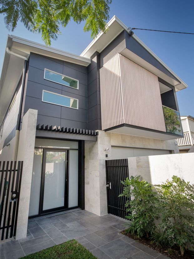Street-level exterior view of a contemporary multi-storey residence. The facade combines dark grey panel cladding, pale textured masonry, white roof and trim elements, a prominent vertically ribbed upper feature wall, black metal gates and detailing, and large glazed openings. A tiled entry path, small landscaped garden, and projecting upper-level form are visible.