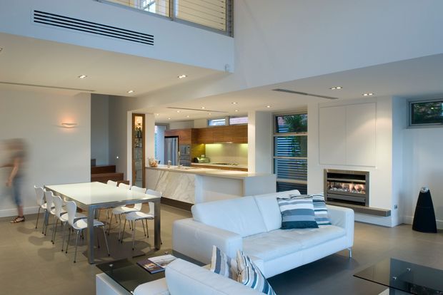 Bright open-plan contemporary interior combining a living room, dining area, and kitchen. The space features a white sectional sofa, glass coffee table, white dining table with sculptural chairs, marble-look kitchen island and splashback, warm timber cabinetry, recessed lighting, a linear fireplace, and a double-height void with upper-level glazing and ventilation grilles.