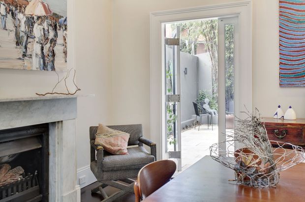 Refined traditional living and dining area with a marble fireplace, framed artwork, upholstered armchair, timber dining table, and a glazed door opening to a small courtyard. The room combines warm timber furniture with pale neutral walls and classic architectural trim.