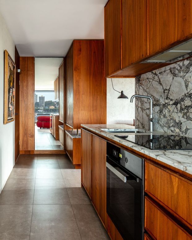 A narrow galley kitchen viewed toward a glazed living area, with extensive warm-toned timber cabinetry, a veined marble backsplash and benchtop, integrated oven and cooktop, stainless steel tap and sink, and large grey floor tiles. A red sofa and high-rise urban outlook are visible in the distance.