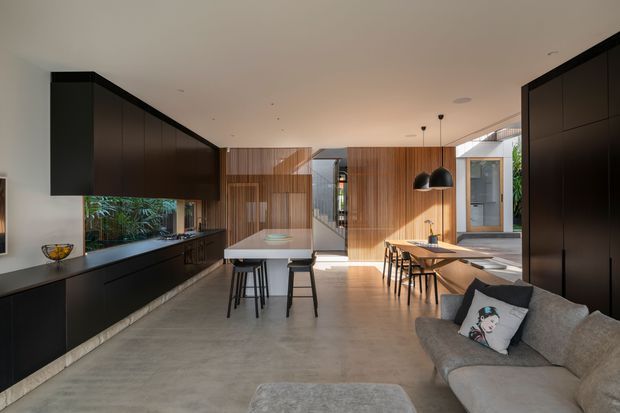 Open-plan contemporary kitchen and dining area with a connected living zone. Dark full-height cabinetry lines the left and right sides, while warm timber wall panelling forms the central backdrop. A pale stone island with stools sits in the kitchen, and a timber dining table with black chairs is positioned near large glazed openings. Polished concrete flooring, black pendant lights, and abundant natural light create a refined, modern interior.