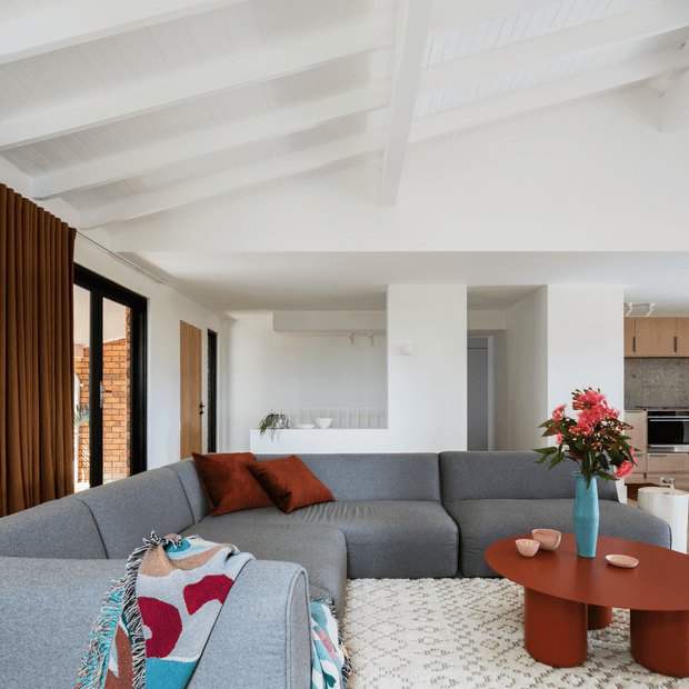 Bright open-plan living room with a large grey sectional sofa, rust-coloured cushions, patterned throw, round terracotta coffee table, and a blue vase with flowers. White walls and ceiling beams create an airy interior, while the rear includes a compact kitchen with timber cabinetry and a speckled grey splashback. Full-height glazing with brown curtains opens toward a brick exterior.
