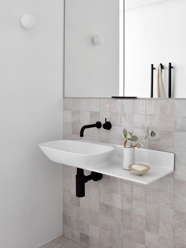 Minimalist contemporary bathroom with a wall-mounted white basin and integrated shelf, matte black wall-mounted tapware and exposed plumbing, pale square tiled wall, large frameless mirror, and simple spherical wall lights. Neutral white and light grey finishes create a clean, refined aesthetic.