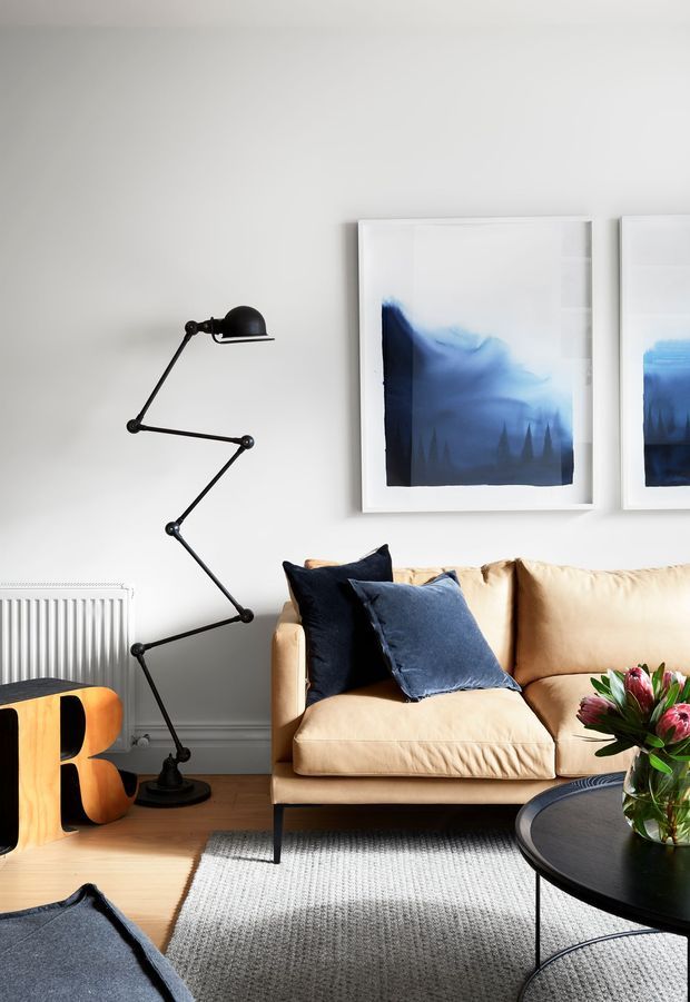 A bright, carefully styled living room vignette with a tan upholstered sofa, navy and blue cushions, a black articulated floor lamp, framed blue-toned artwork, a round black coffee table, light timber flooring, and a pale textured rug. A white radiator and sculptural wooden side object are also visible. The room has a clean contemporary aesthetic with strong navy-and-neutral contrast.