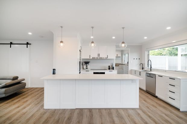 Bright, open-plan white kitchen with a large paneled island, extensive white cabinetry, light timber-look flooring, pendant lights, stainless-steel appliances, and a wide window above the sink. A partial living area and barn-style sliding door are visible at the left.