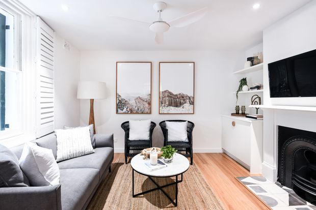 Bright, compact living room with white walls, light timber flooring, a grey sofa, two black accent chairs, a round coffee table, woven rug, built-in white shelving and cabinetry, and a traditional dark fireplace with a wall-mounted television above it. Plantation shutters, framed artwork, recessed lighting, and a ceiling fan complete the clean, relaxed interior.