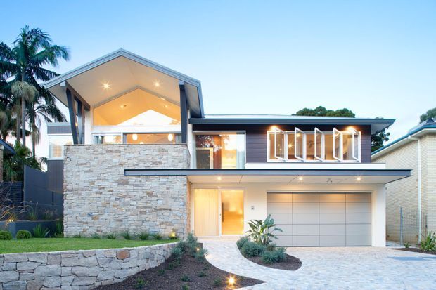 Contemporary two-storey residential facade at dusk, featuring a broad low-pitched roof, dramatic open gable with exposed soffit, extensive glazing, a projecting upper-level balcony or terrace, pale stacked-stone wall cladding, and an integrated double garage. A curved light-paved path and landscaped planting lead to the entry, with tropical palms visible at the side.