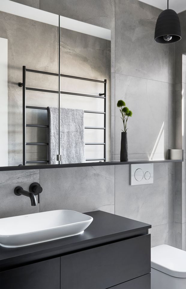 Contemporary bathroom featuring large-format grey concrete-look wall tiles, a frameless mirrored wall, matte black wall-mounted tapware, a black heated towel rail, and a dark floating vanity with a white vessel basin. A black pendant light, minimalist accessories, and a concealed cistern toilet complete the refined monochromatic scheme.