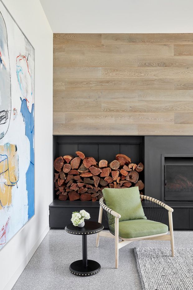 A refined living room corner featuring a black built-in fireplace with an open firewood storage niche, pale timber wall cladding above the fireplace, and a contemporary armchair in muted green upholstery. A small black side table, abstract artwork, textured rug, and light grey carpet complete the calm, modern interior.