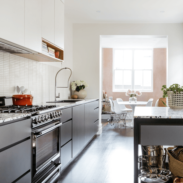A bright, refined galley kitchen with dark grey flat-panel cabinetry, a large stainless-steel range cooker, pale horizontal tile splashback, and light stone benchtops. A dining area is visible beyond the kitchen, with a white table and chairs positioned in front of a large window. The room features polished dark timber flooring, white walls, integrated upper cabinetry, and a black island or peninsula with open storage and cookware beneath.