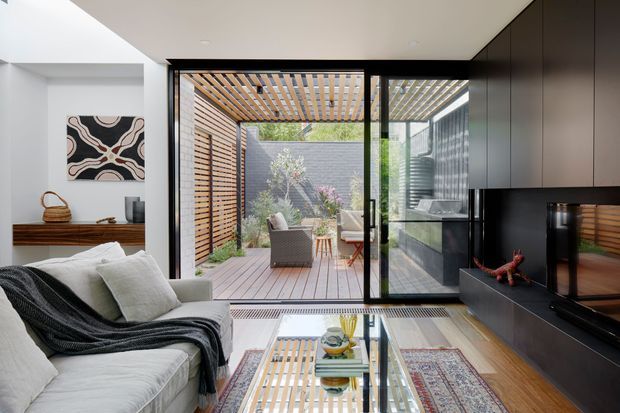 Bright contemporary living room opening directly to a private courtyard through large sliding glass doors. The room features a light sofa with dark throw, built-in dark cabinetry and media wall, timber flooring, a glass coffee table, and a timber-slatted pergola and screen visible outside. Strong indoor–outdoor connection with abundant natural light.