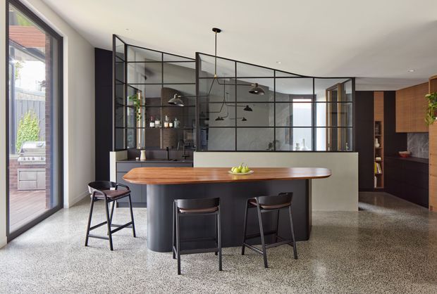 A sophisticated open-plan kitchen with a large island featuring a rounded timber benchtop and dark curved cabinetry, surrounded by bar stools. A black metal-framed glass partition separates the kitchen from an adjoining living or dining area. The space has polished terrazzo-style flooring, dark joinery, pendant lighting, and expansive glazing with an outdoor grill visible beyond.