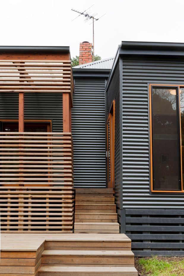 Exterior view of a compact contemporary house with dark corrugated metal cladding, timber-framed windows, extensive horizontal timber privacy screening and decking, and a stepped timber entry staircase. A brick chimney and corrugated pitched roof are visible above the adjoining volumes.