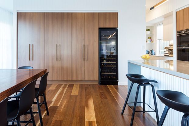 Bright contemporary open-plan kitchen and dining area with extensive timber cabinetry, a built-in wine fridge, timber flooring, a stone island bench with a vertically fluted front, black bar stools, and a dark timber dining table.