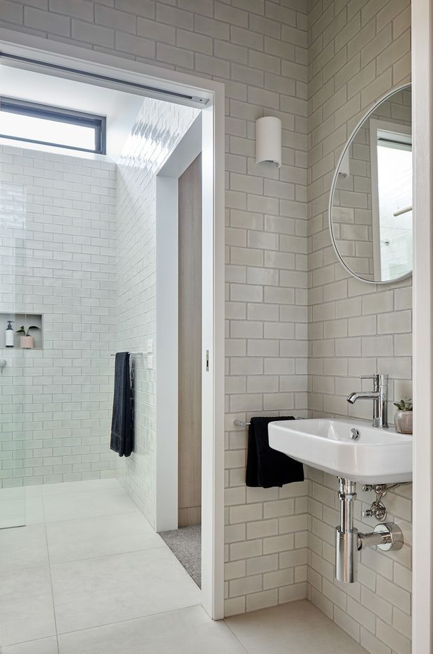 Bright, compact contemporary bathroom with pale grey-white brick-pattern wall tiles, a walk-in tiled shower visible through an open doorway, a wall-mounted white basin with exposed chrome plumbing, an oval wall mirror, and minimalist cylindrical wall lighting. The space is clean and light-filled, with a small high window and subtle timber-toned cabinetry visible in the shower area.