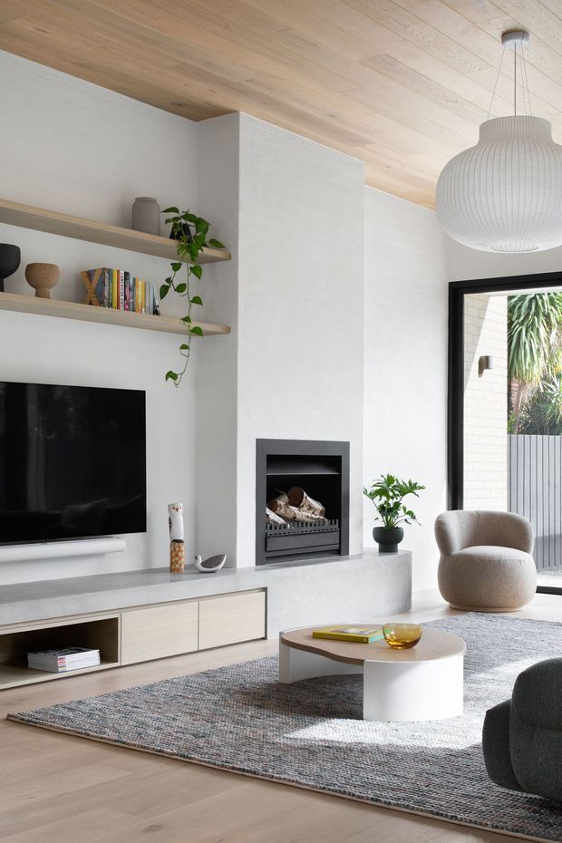 Bright contemporary living room with a pale timber-lined ceiling, white textured walls, built-in open shelving, a wall-mounted television, and a minimalist fireplace integrated into a central white chimney breast. A long low grey built-in media cabinet runs beneath the television and fireplace. The room includes a rounded upholstered armchair, sculptural white coffee table, dark lounge chair, large textured rug, indoor plant, and oversized ribbed pendant light. Full-height black-framed glazing opens to an exterior courtyard or garden area.
