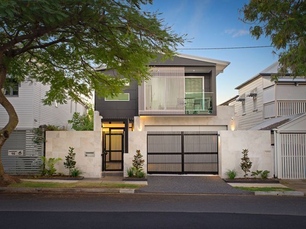 Street-facing view of a contemporary multi-storey urban residence with a pale stone or rendered lower facade, dark upper wall panels, vertical screening, glass balcony, flat projecting roof and black metal vehicle and pedestrian gates. Narrow landscaped planting borders the driveway and frontage, with neighbouring attached or closely spaced homes visible on both sides.