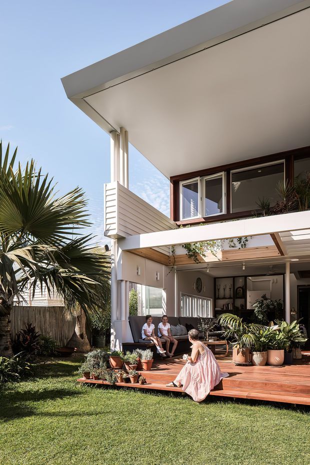 Exterior view of a contemporary two-storey home opening onto a timber deck and landscaped lawn. The home features a broad flat roof and deep covered outdoor living area, white horizontal cladding, dark timber-framed glazing, exposed timber soffit detailing, and extensive potted planting. People are seated on the deck, with a palm-filled garden in the foreground.
