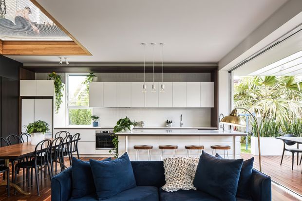 Open-plan contemporary kitchen, dining, and living area with a dark navy sofa in the foreground, timber dining table and black spindle-back chairs to the left, and a bright white kitchen with a central island and timber stools beyond. Large glazed openings connect the room to a timber deck and lush outdoor planting. A roof skylight and dark timber-toned cabinetry create contrast against the light walls and ceiling.
