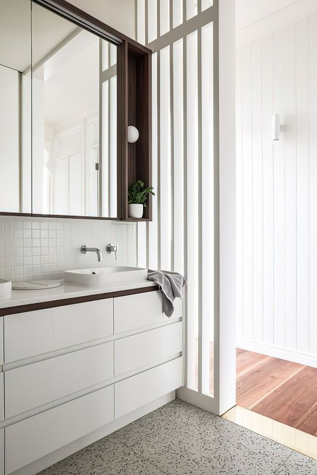 Bright contemporary bathroom vanity with a floating white drawer unit, integrated white basin, wall-mounted chrome tapware, and a small square white-tile splashback. A dark timber-framed mirrored cabinet with open shelving sits above the vanity. Vertical white wall panelling and a tall white-framed glazed partition or door bring strong natural light into the room, with terrazzo-style flooring and timber flooring visible beyond the threshold.