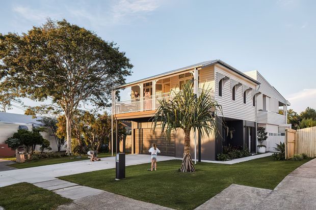 Exterior view of a contemporary two-storey residence with light horizontal weatherboard cladding, a pitched roof, covered upper-level balcony, and a ground-floor garage or covered parking area. The home is set within a landscaped suburban site with a manicured lawn, concrete paths and driveway, mature trees, and tropical-style planting.