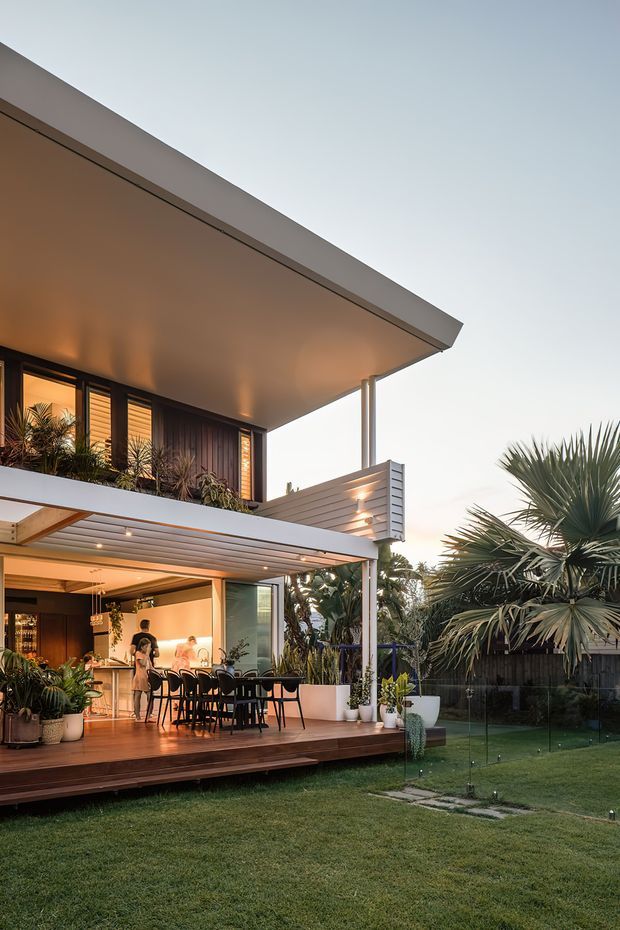 Exterior view of a contemporary two-storey home at dusk, focused on a covered outdoor dining deck and open-plan dining/living area. The house features broad flat roof planes, slim posts, extensive glazing, horizontal cladding, timber-lined soffits, warm interior lighting, and a lush tropical garden with a lawn and palms.