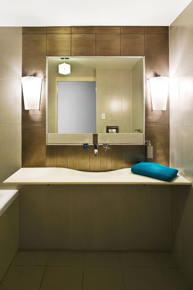 Compact contemporary bathroom vanity with a wide white integrated basin and double wall-mounted faucets beneath a large mirrored cabinet. Dark timber-look wall panelling frames the mirror, while beige tiled walls and floor create a warm neutral scheme. Vertical wall sconces flank the mirror, and a bright turquoise folded towel provides the main accent.