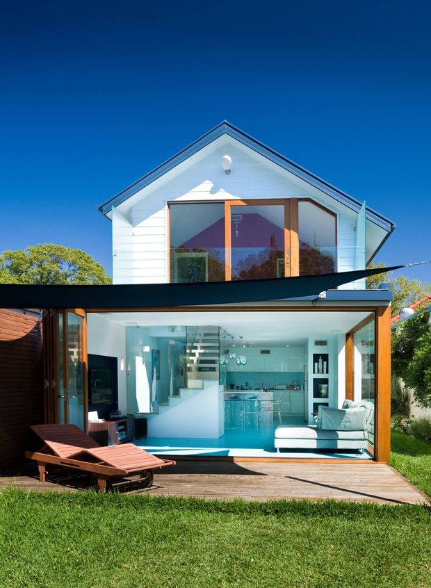 Bright exterior view of a compact two-storey house with a white gabled upper facade, extensive timber-framed glazing, and an open-plan ground-floor living area visible through fully opened glass doors. The interior includes a contemporary kitchen, glass stair enclosure, and pale sectional sofa. A timber deck with lounge chairs, a dark shade sail, and a lawn occupy the foreground. The image has a crisp, sunny, indoor-outdoor character.