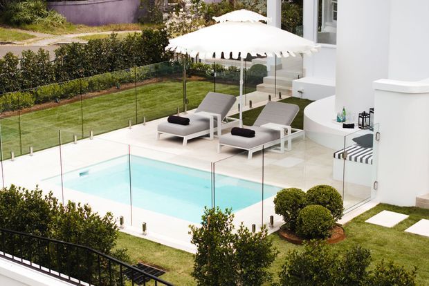 A refined outdoor pool area beside a white contemporary residence, featuring a pale turquoise swimming pool enclosed by frameless glass safety fencing, white paving, sculpted topiary, manicured lawn, white sun loungers, and a large white patio umbrella. A curved white architectural wall and outdoor seating area frame the right side.