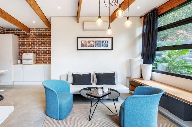 A bright, compact contemporary living room with a white sofa, two vivid blue armchairs, a round black coffee table, and a long timber window seat beneath large black-framed glazing. Exposed timber ceiling beams, exposed red brickwork, polished concrete flooring, pendant lights, and white built-in cabinetry create an industrial-meets-contemporary interior. A small work area with cabinetry and a printer is visible at the rear.